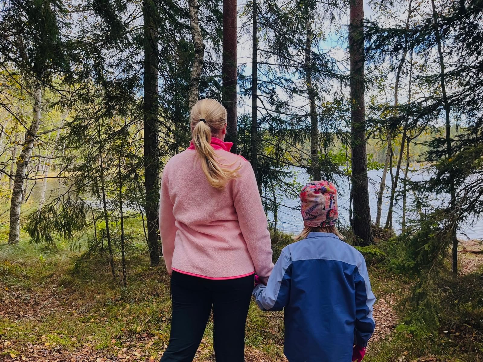 Lastenvahti ja lapsi kädet käsin järvimaisemalla metsän reunassa